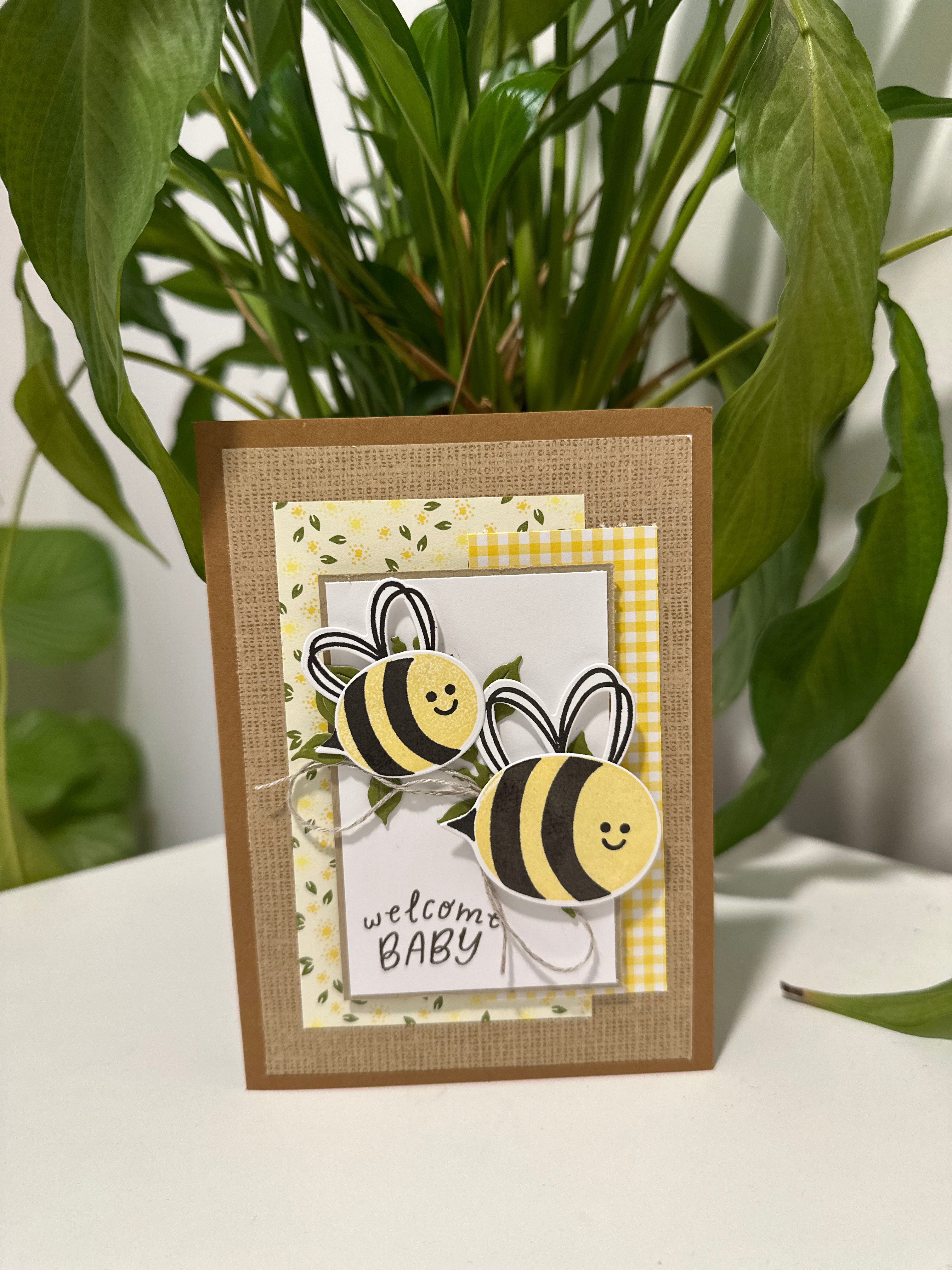 Greeting card with bee design and 'welcome baby' text, placed on a surface with green leaves in the background.