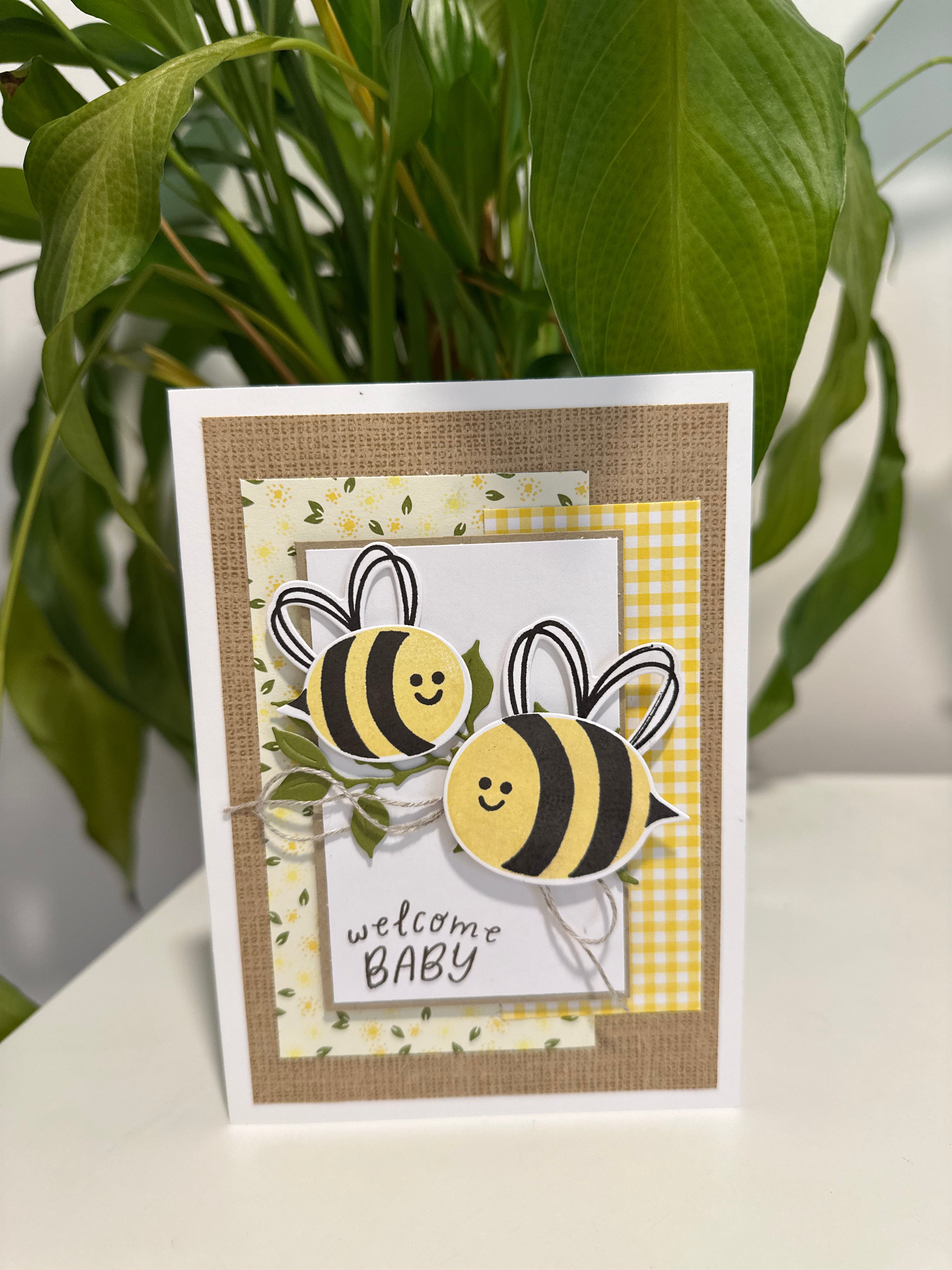 Greeting card with bee designs and 'welcome baby' text, placed on a surface with a plant in the background.