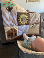 A baby sensory play mat with whimsical appliques including a sun, tree and barn with a baby in a bouncinette looking at it