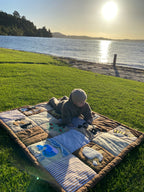 A baby lies on a sensory play mat  on a green grassy lawn next to a lake at sunset, with the sun reflecting off the water in the background.