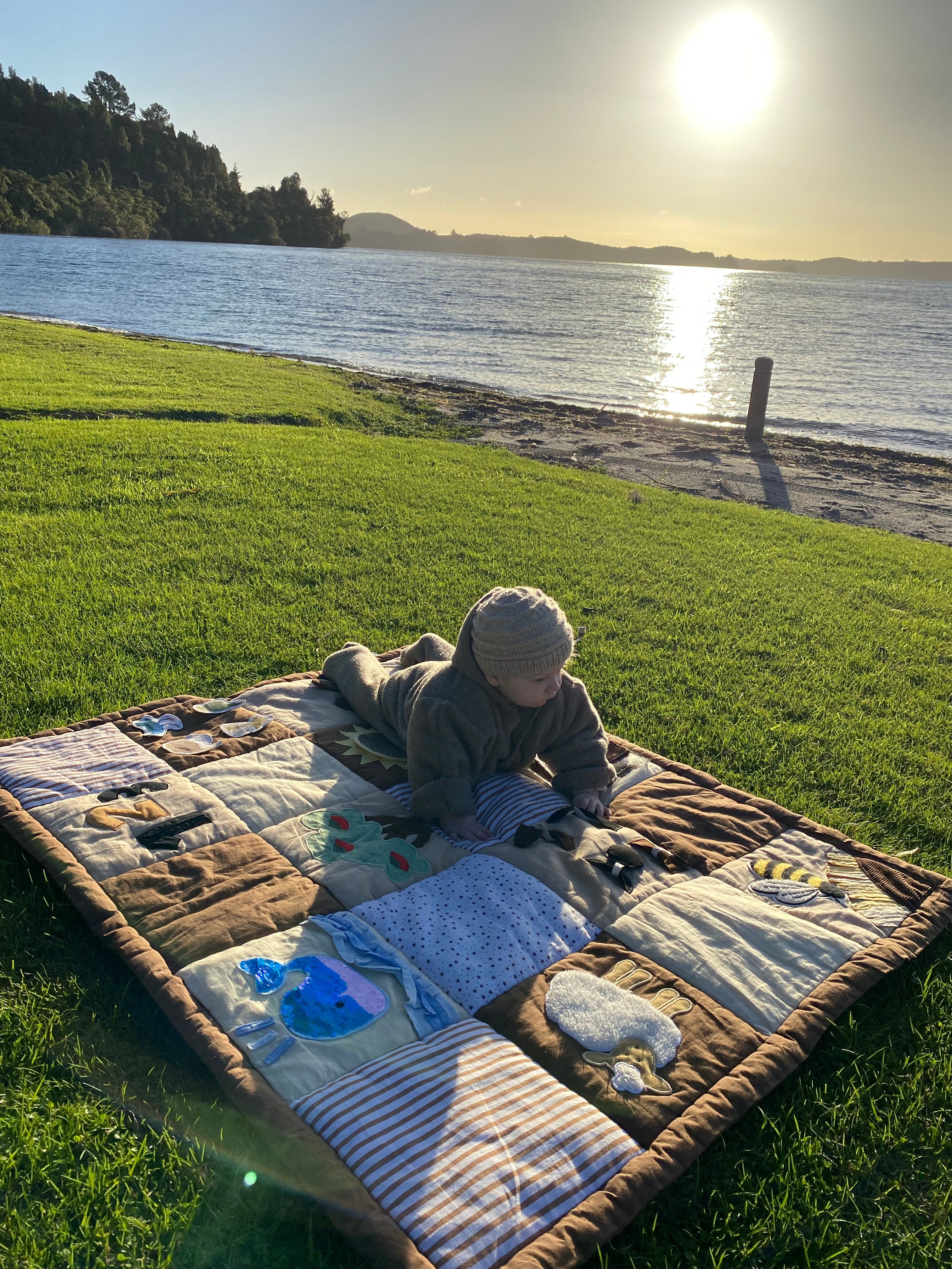 A baby lies on a sensory play mat  on a green grassy lawn next to a lake at sunset, with the sun reflecting off the water in the background.