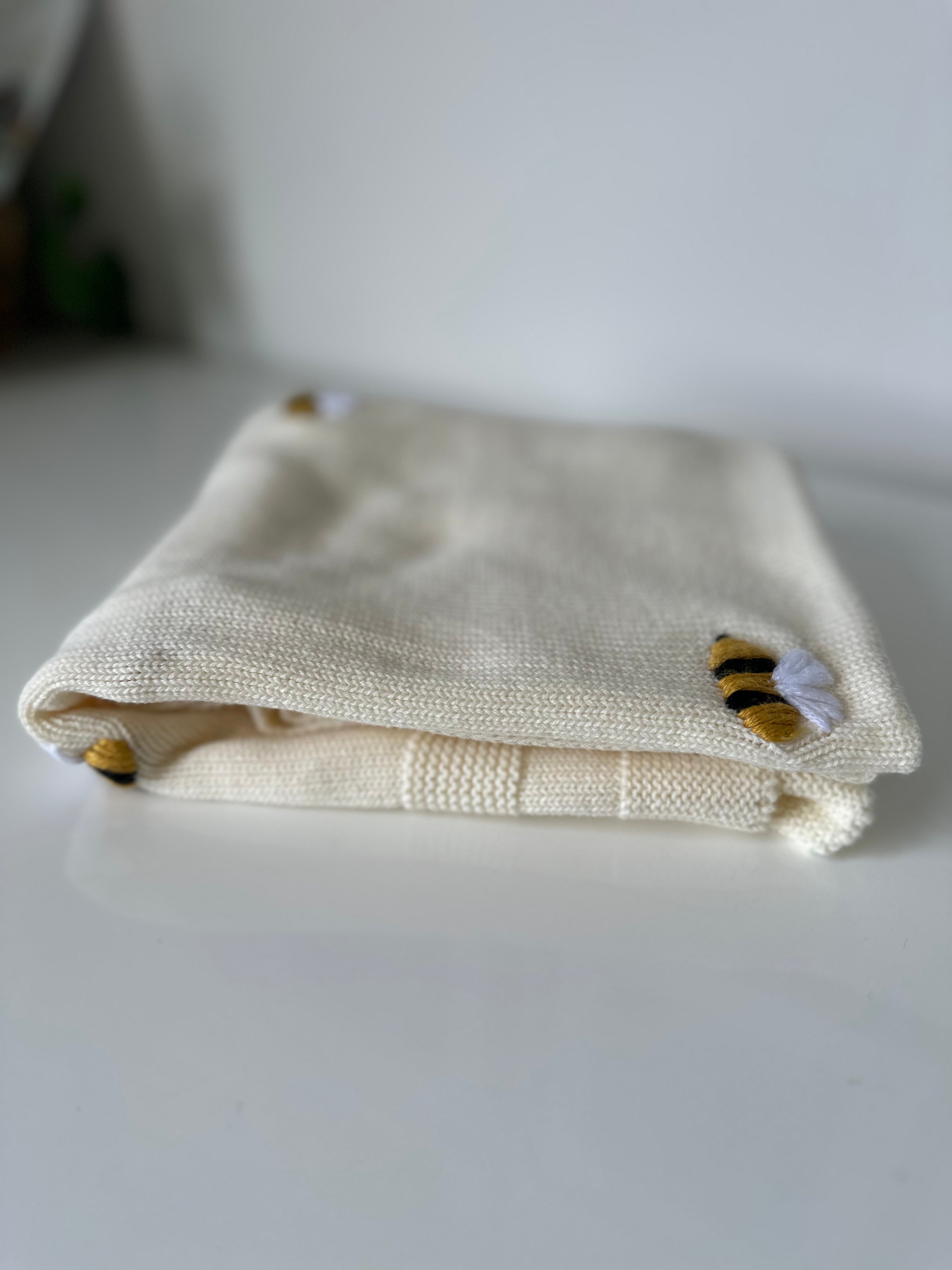 Folded cream knit baby blanket with bee designs on a white surface