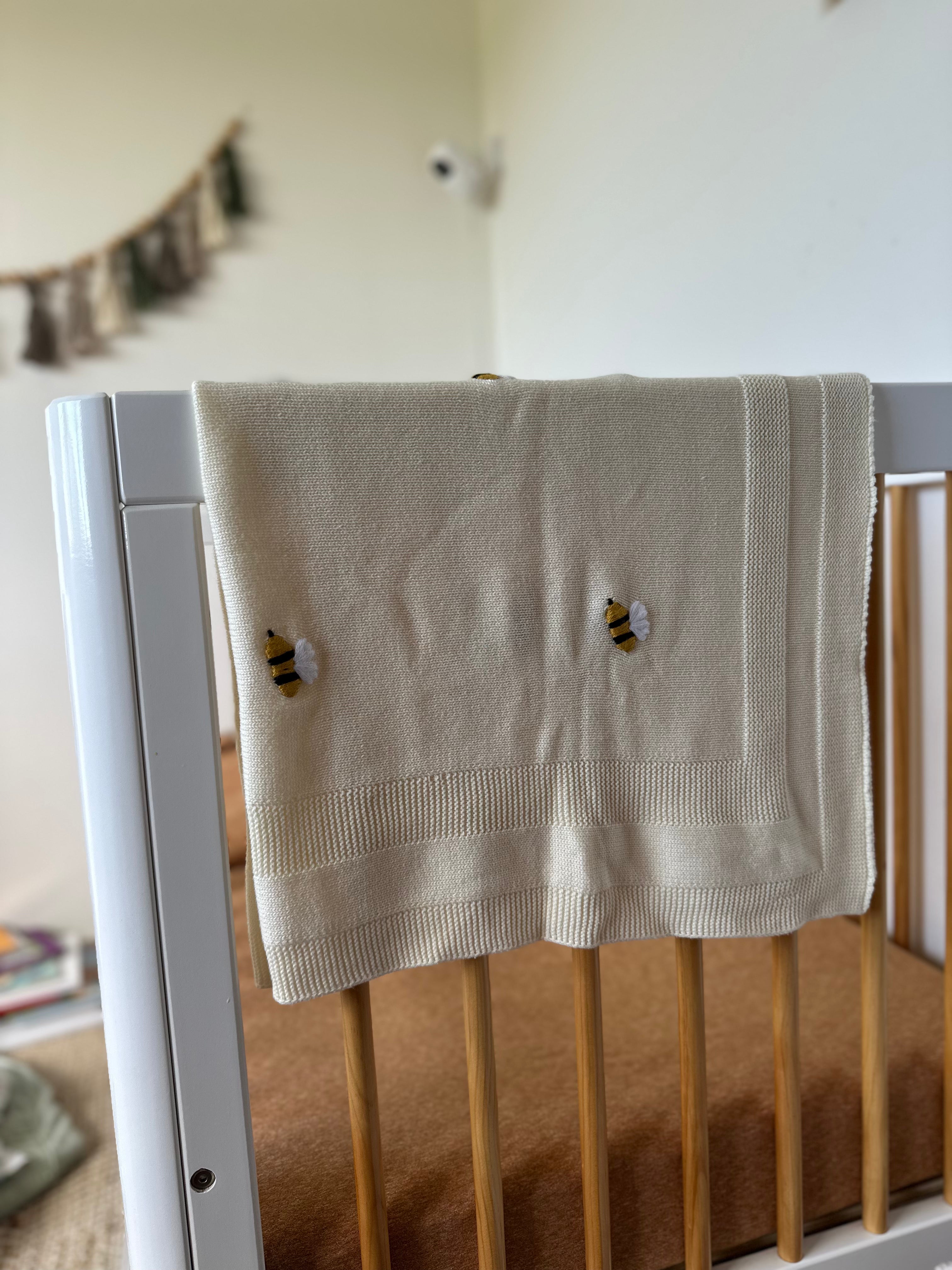 cream baby blanket with bee patterns draped over a crib/cot in a nursery.