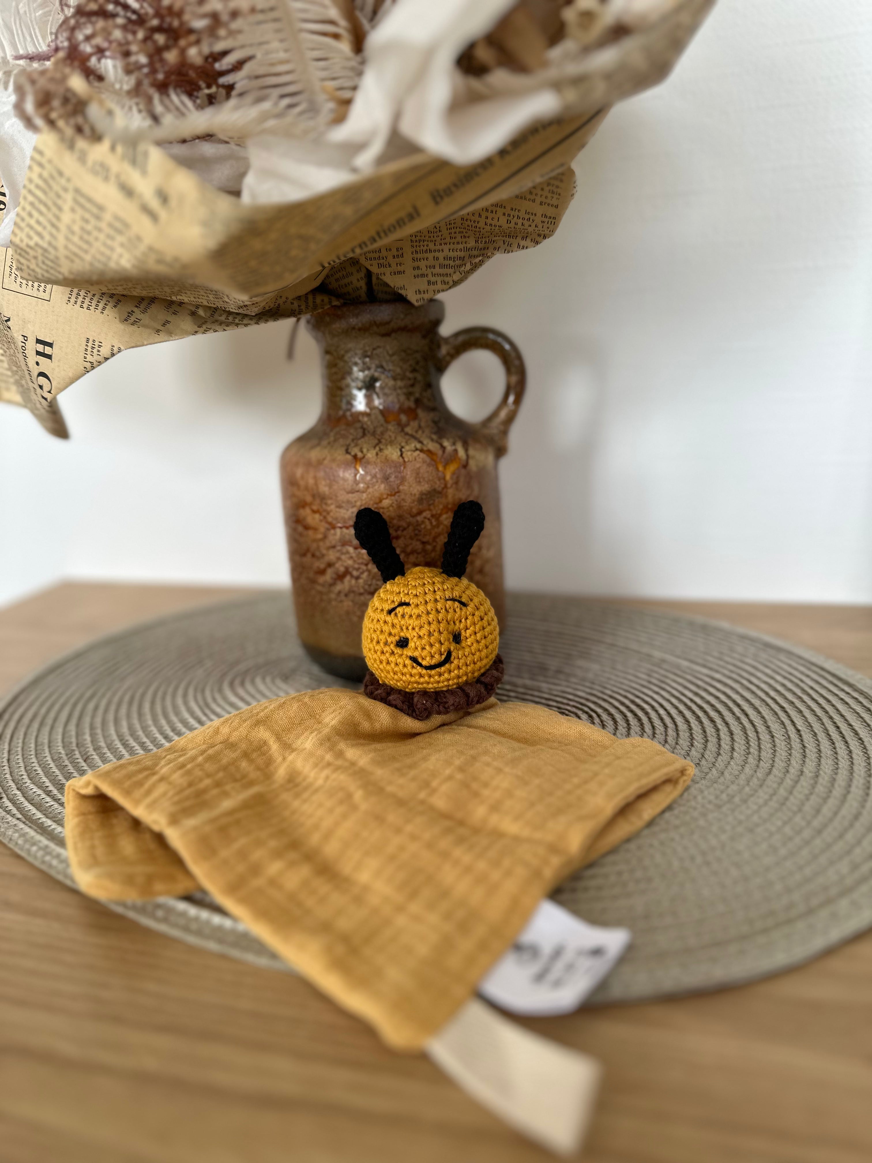 Small crocheted bee comforter toy on a yellow material with a vase and newspaper in the background.