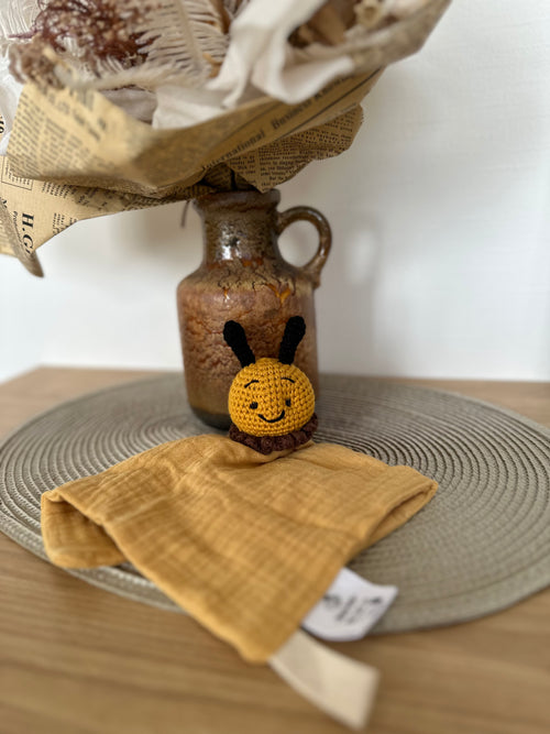 Small crocheted bee comforter toy on a yellow material with a vase and newspaper in the background.