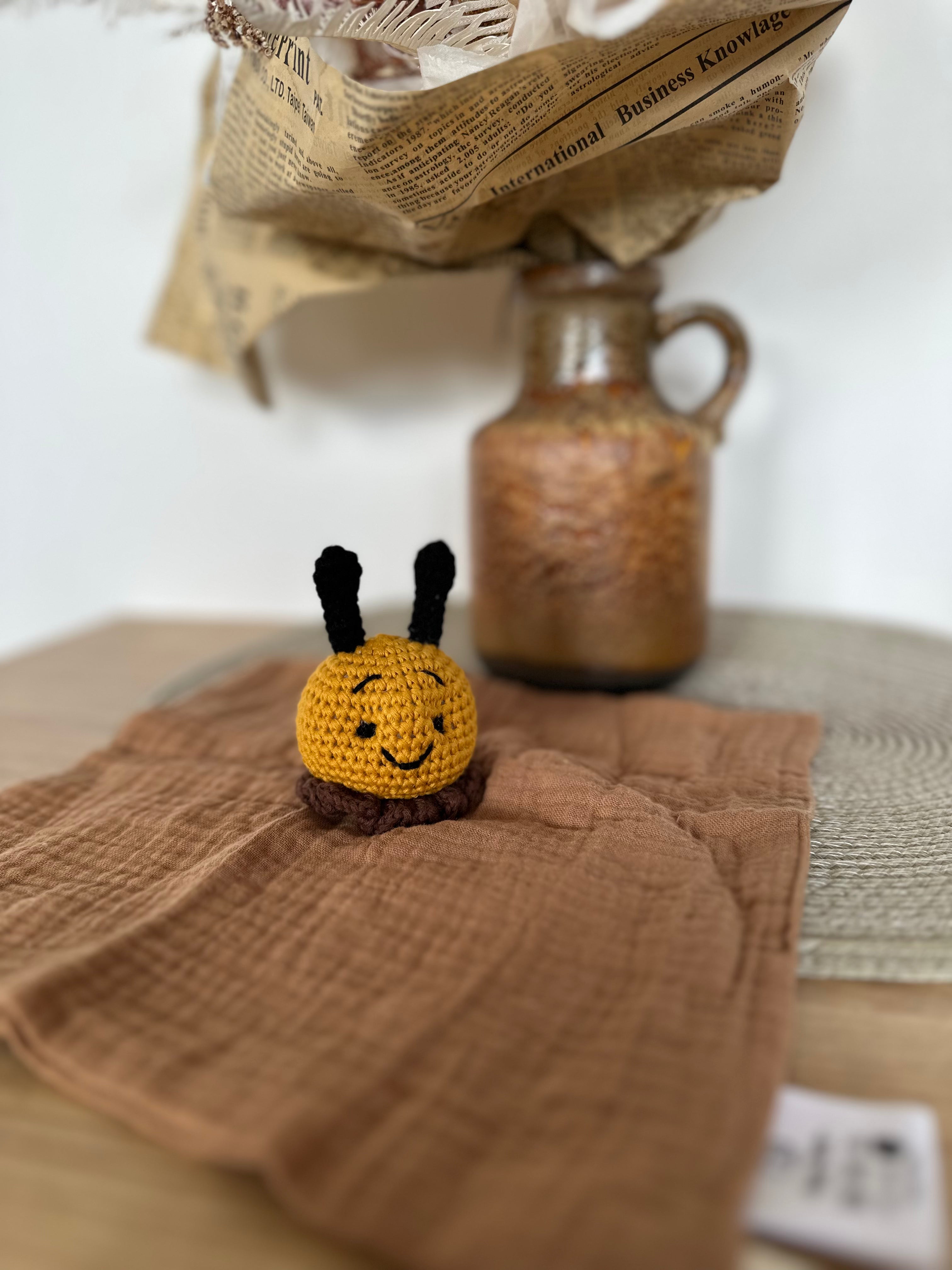 Small crochet bee toy on a brown textured fabric with a vase and newspaper in the background.