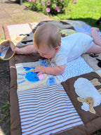 Baby playing on a colorful play mat outdoors looking at the sequined whale and holding the circle mirror