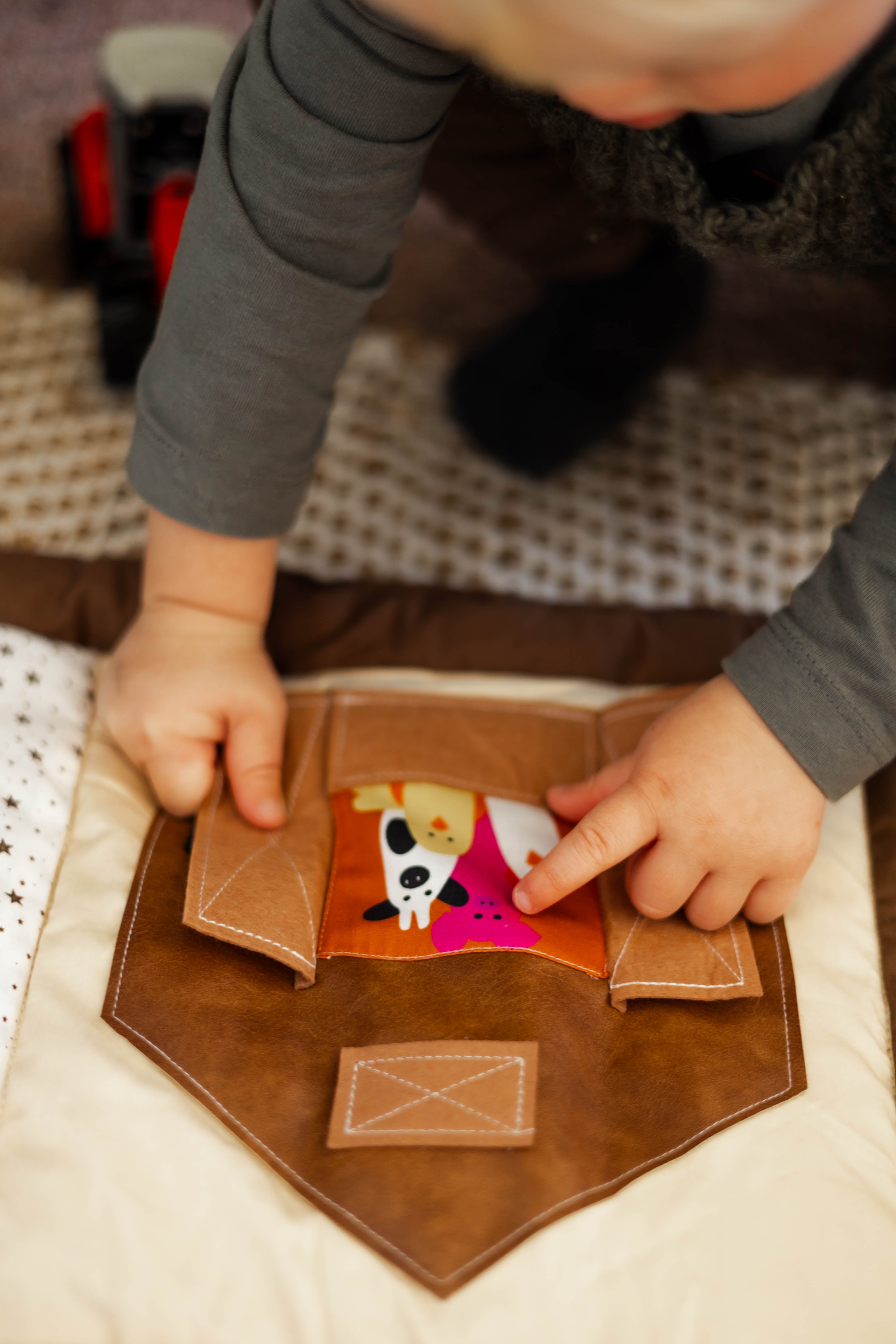 Child playing with a brown leather material doors featured on a baby play mat with cartoon characters on a textured surface.