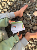 A child sitting on a bed of small gray rocks holding Nosey Toesie Wild Wander Cards